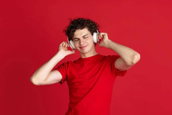 Retrato recortado de un joven en tela casual escuchando música en auriculares aislados sobre fondo rojo —  Fotos de Stock