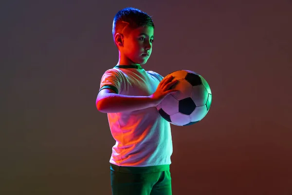 Retrato recortado de niño, niño, entrenamiento de jugador de fútbol aislado sobre fondo degradado en luz de neón. — Foto de Stock