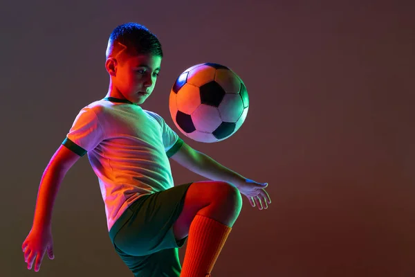 Ausgeschnittenes Porträt von Junge, Kind, Fußballspieler beim Training, kickender Ball mit Knie isoliert über Steigungshintergrund im Neonlicht. — Stockfoto