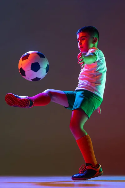 Ausgeschnittenes Porträt von Junge, Kind, Fußballspieler beim Training mit Ball isoliert über Gradienten Hintergrund in Neonlicht. — Stockfoto