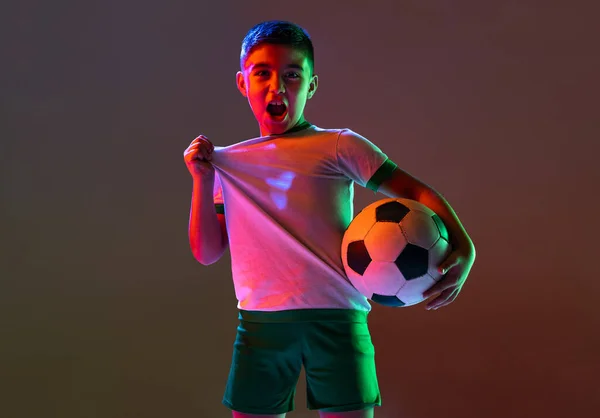 Retrato recortado de niño, niño, jugador de fútbol posando aislado sobre fondo degradado en luz de neón — Foto de Stock