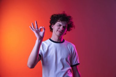 Beyaz tişörtlü genç bir adamın kesilmiş portresi. Hoş bir jest. Neon ışıklarıyla izole edilmiş pembe turuncu bir arkaplan.