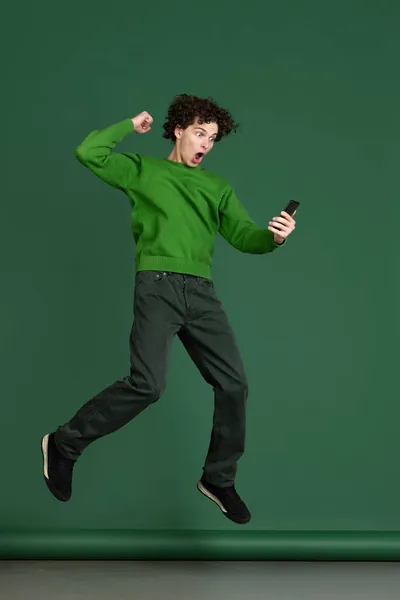 Retrato de comprimento total de jovem em suéter verde olhando para o telefone e pulando isolado sobre fundo verde escuro. Notícias animadas — Fotografia de Stock