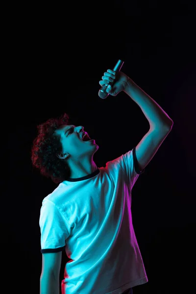 Vágott portré fiatal férfi fehér pólóban kifejezetten énekel mikrofonban elszigetelt fekete háttér neon fények — Stock Fotó