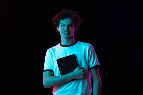 흰색 티셔츠를 입고 네온등에 검은 배경으로 분리 된 태블릿이 있는 젊은 남성의 사진이 찍혀 있다. — 스톡 사진