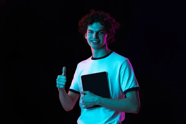 Vágott portré fiatal férfi fehér pólóban tabletta mutatja, mint a gesztus elszigetelt fekete háttér neon fények — Stock Fotó