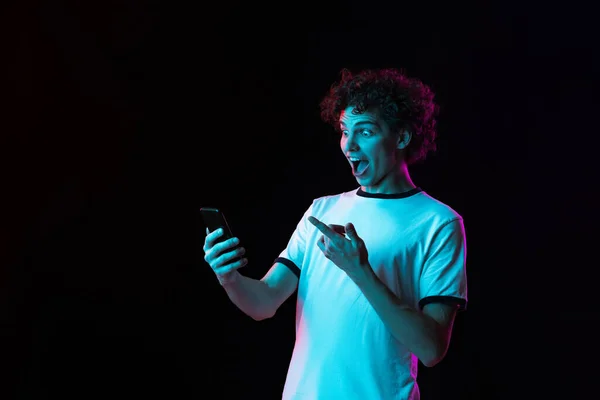 Vágott portré egy fehér pólós fiatalemberről, akinek a telefonja fekete háttérrel van elszigetelve a neonfényektől. Boldog és izgatott nézés — Stock Fotó