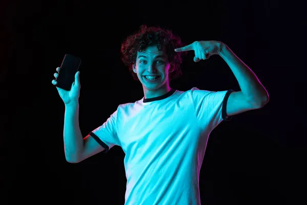 Vágott portré egy fehér pólós fiatalemberről, aki a fekete háttér felett elszigetelt telefonképernyőre mutat neonfényben. Boldog és izgatott nézés — Stock Fotó