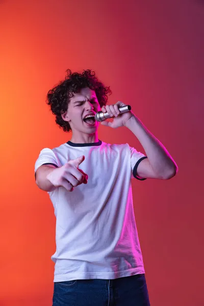 Ausgeschnittenes Porträt eines jungen Mannes in weißem T-Shirt, der in Mikrofon singt, isoliert über einem Gradienten in rosa-orangefarbenem Hintergrund in Neonlichtern — Stockfoto
