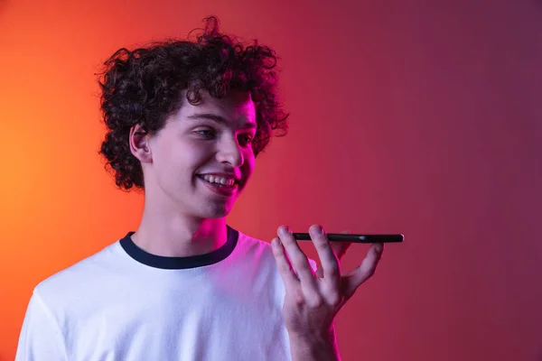 Ausgeschnittenes Porträt eines jungen Mannes in weißem T-Shirt, der eine Sprachnachricht am Telefon aufzeichnet, isoliert über einem Gradienten in rosa-orangefarbenem Hintergrund im Neonlicht — Stockfoto