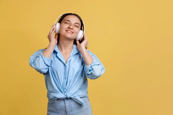 노란색 배경으로 고립된 헤드폰으로 음악을 듣고 있는 아름다운 파란 셔츠를 입은 젊은 여성의 사진이 찍혀 있다 — 스톡 사진