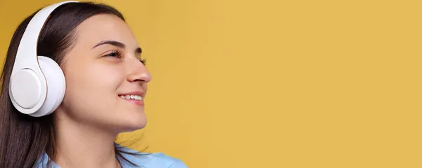 Gros plan portrait de jeune belle femme souriante en chemise bleue écoutant de la musique dans des écouteurs isolés sur fond jaune. FLyer — Photo