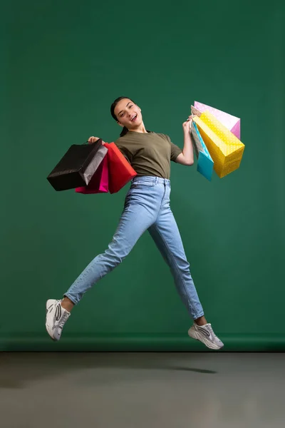 Ritratto a figura intera di giovane donna felice in tessuto casual che salta con borse della spesa isolate su sfondo verde — Foto Stock