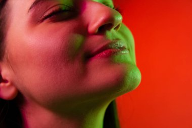 Kapalı gözlerle poz veren güzel bir kadının yakın plan portresi neon ışıklarla izole edilmiş kırmızı arka plan üzerinde.