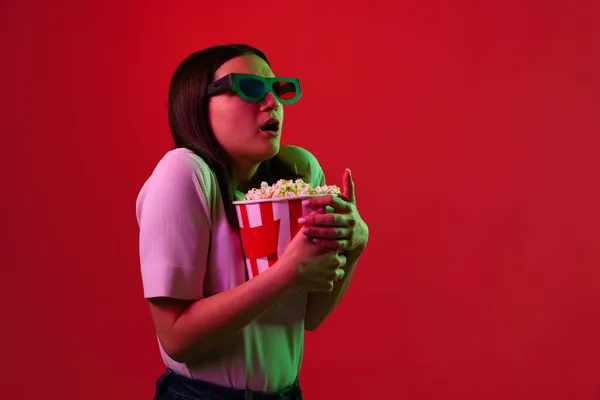 Retrato recortado de una joven hermosa mujer en camiseta blanca y gafas 3D comiendo palomitas aisladas sobre fondo rojo en luces de neón —  Fotos de Stock