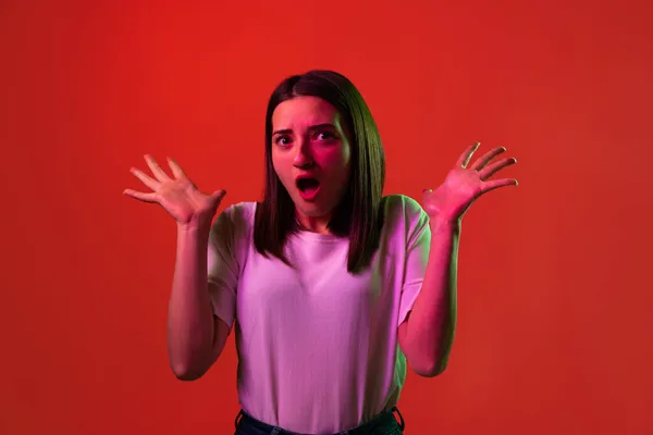 Retrato recortado de jovem mulher bonita em branco T-shirt espalhando as mãos em choque isolado sobre fundo vermelho em luzes de néon — Fotografia de Stock