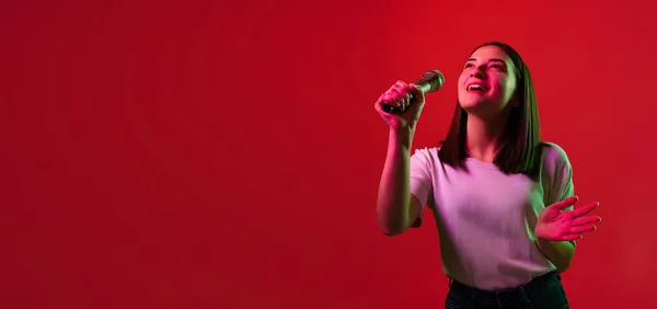 Retrato recortado de jovem mulher bonita em branco T-shirt cantando em microfone isolado sobre fundo vermelho em luzes de néon. Folheto — Fotografia de Stock