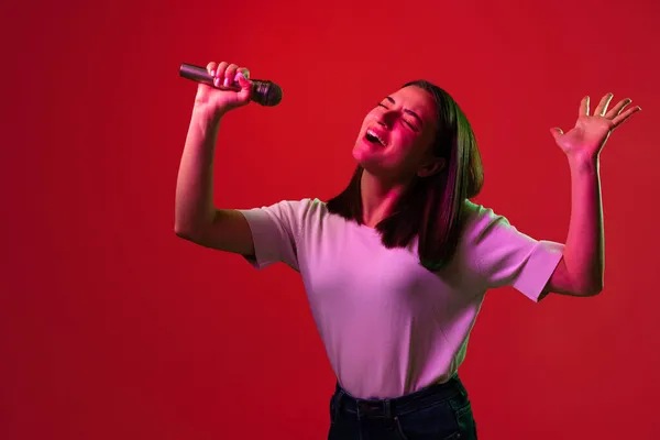흰색 티셔츠를 입은 젊고 아름다운 여성 이 네온 조명을 받은 붉은 배경에서 고립된 마이크로 크게 노래하는 모습을 그린 사진 — 스톡 사진
