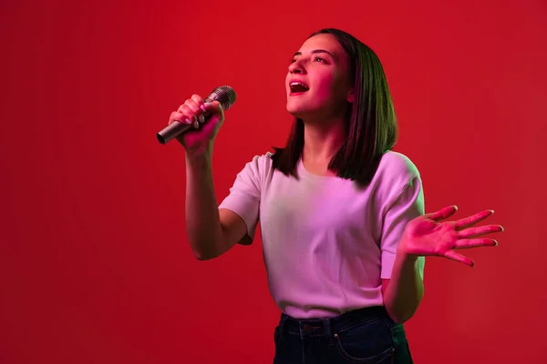 Vágott portré fiatal gyönyörű nő fehér pólóban énekel mikrofonban elszigetelt piros háttér neon fények — Stock Fotó