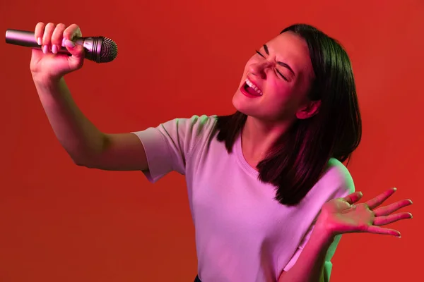 Gesneden portret van jonge mooie vrouw in wit T-shirt expressief zingen in microfoon geïsoleerd over rode achtergrond in neon lichten — Stockfoto