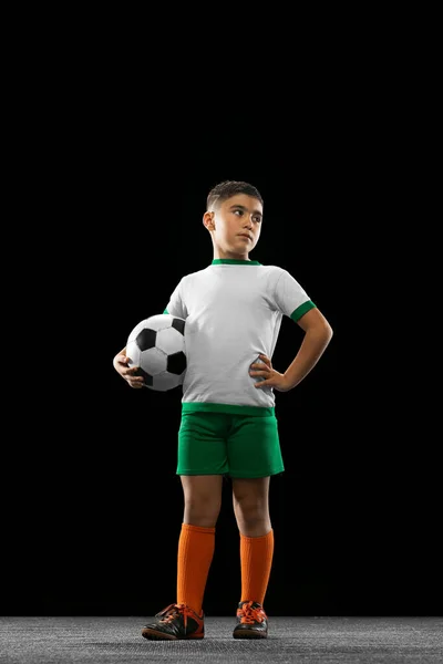 Pełnowymiarowy portret chłopca, dziecka, piłkarza w mundurze pozującego z piłką odizolowaną na czarnym tle. Obrońca — Zdjęcie stockowe