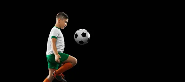 Retrato completo de menino, criança, jogador de futebol em treinamento uniforme, chutando bola com joelho isolado sobre fundo preto. Folheto — Fotografia de Stock
