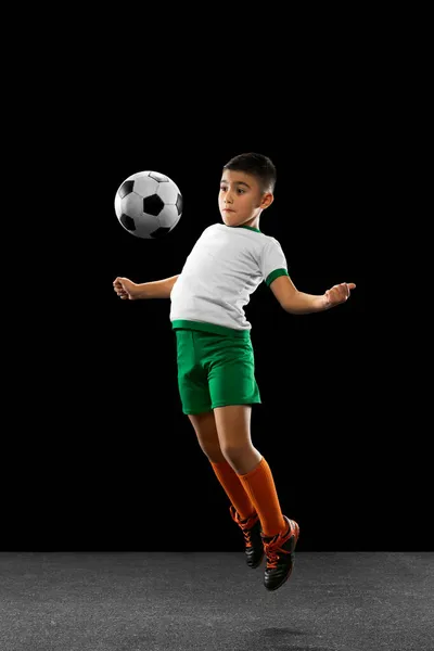 Retrato completo de niño, niño, jugador de fútbol en entrenamiento uniforme, pateando la pelota con el pecho en un salto aislado sobre fondo negro. —  Fotos de Stock