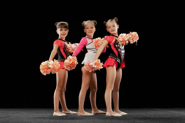 Retrato completo de três meninas, líderes de torcida treinando em trajes de palco com pom-pons isolados sobre fundo preto — Fotografia de Stock