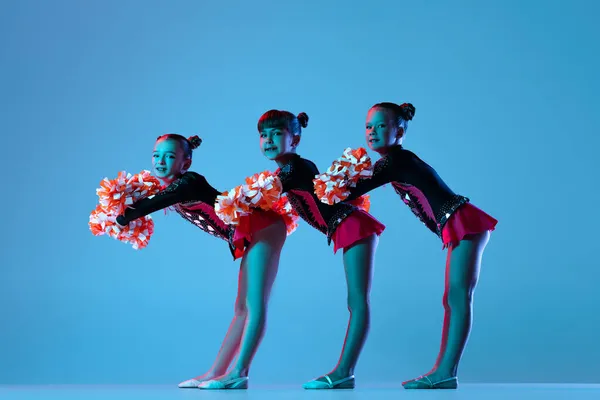 Ganztägiges Porträt von drei Mädchen, Cheerleadern beim Training in bunten Kostümen mit Bommelmützen vor blauem Hintergrund in Neonlicht — Stockfoto
