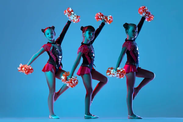Full-length portret van drie meisjes, cheerleaders training in kleurrijke kostuums met pompons geïsoleerd over blauwe achtergrond in neon lichten — Stockfoto