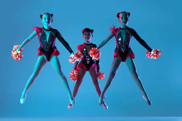 Full-length portret van drie meisjes, cheerleaders training in kleurrijke kostuums met pompons geïsoleerd over blauwe achtergrond in neon lichten — Stockfoto