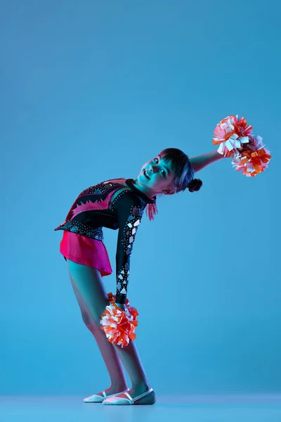 Full-length portret van mooi meisje, cheerleader training, optreden in helder bodysuit met pompons geïsoleerd over blauwe achtergrond in neon lichten — Stockfoto