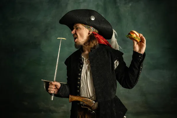 Retrato de un hombre brutal, pirata medeival sosteniendo hamburguesa aislada sobre fondo oscuro. — Foto de Stock