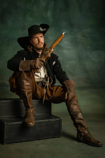 Обрезанный портрет жестокого пугающего человека, медеивального пирата в винтажном костюме с мечом и пистолетом, сидящего изолированно на тёмном фоне. — стоковое фото