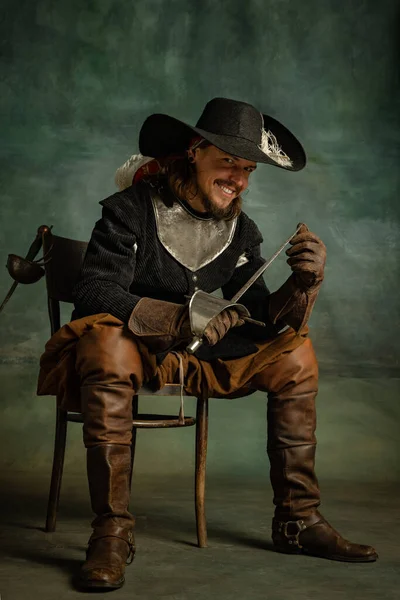 Портрет жестокого ужасающего человека, медейвального пирата в винтажном костюме, сидящего и держащего меч изолированным на тёмном фоне. — стоковое фото