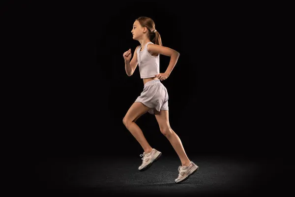 Full-length portrait of girl, teenager, athlete training running isolated over black background. Side view — Stock Photo, Image