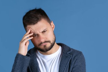 Ciddi görünen, kafaya dokunan yorgun bir adamın mavi arka planda soyutlanmış bir portresi.