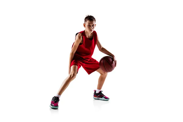 Ganzkörperporträt eines Jungen, Basketballspielers in roter Uniform, der isoliert vor weißem Hintergrund trainiert. Dribbelball — Stockfoto