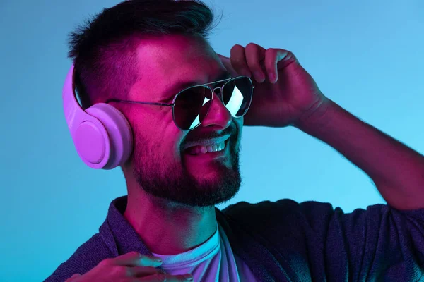 Portrait of man in sunglasses cheerfully listening to music in white headphones isolated over blue background — Stock Photo, Image