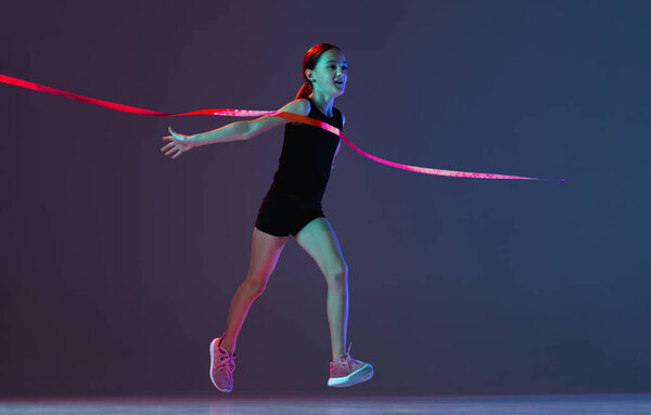 Full-length portrait of teen girl, athlete crossing red ribbon isolated over gradient blue purple background in neon. Winning