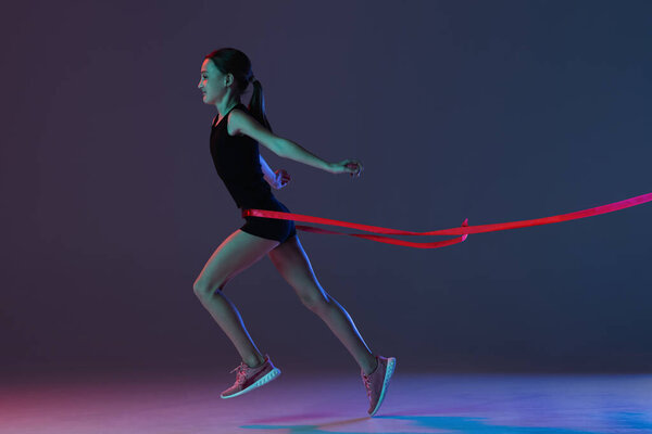 Full-length portrait of teen girl, athlete crossing red ribbon isolated over gradient blue purple background in neon
