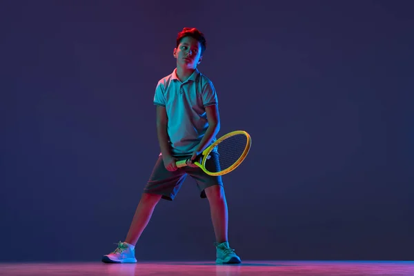 Full-length portrait of boy, tennis player posing isolated over blue background in neon lights — Stock Photo, Image