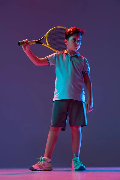 Full-length portrait of sportive boy, tennis player posing isolated over purple background in neon lights — Stock Photo, Image