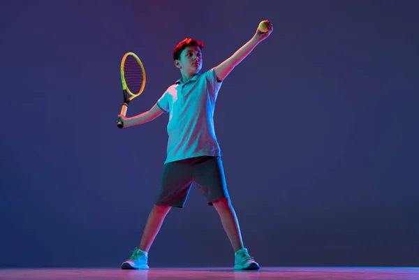 Full-length portrait of boy, tennis player in motion, kicking ball with racket isolated over purple background in neon — Stock Photo, Image