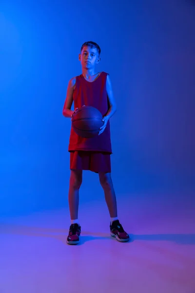 Porträt eines jugendlichen Sportlers, Basketballspieler in Uniform posiert isoliert über einem Gefälle blaulila Hintergrund im Neonlicht — Stockfoto