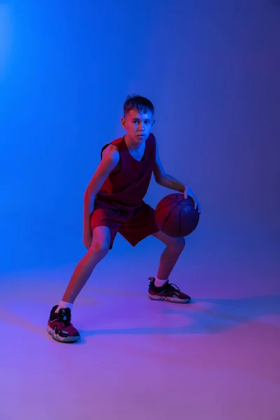 Retrato completo de niño, jugador de baloncesto en movimiento, entrenamiento aislado sobre fondo azul púrpura degradado en luces de neón —  Fotos de Stock