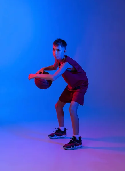 Портрет спортивного мотивированного мальчика в движении, тренировки, игры в баскетбол изолированы на градиентно-синем фиолетовом фоне в неоновых огнях — стоковое фото