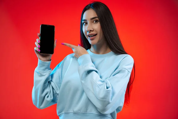 Cropped portrait of young joyful girl pointing at phone screen isolated over red background — Stock Photo, Image