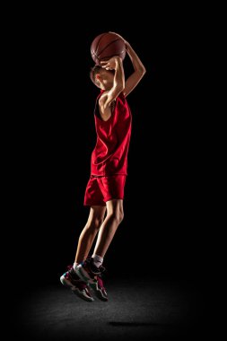 Genç bir çocuğun tam boy portresi, basketbol oyuncusu eğitimi, siyah arka planda izole bir şekilde topu sepetin içine atmak.