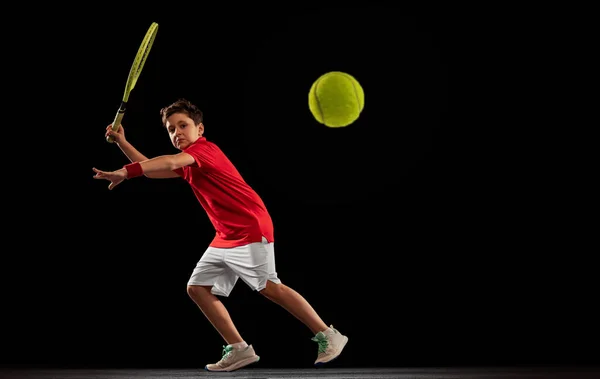 Retrato completo de menino, jogador de tênis em movimento, chutando bola com raquete isolado sobre backgound preto — Fotografia de Stock